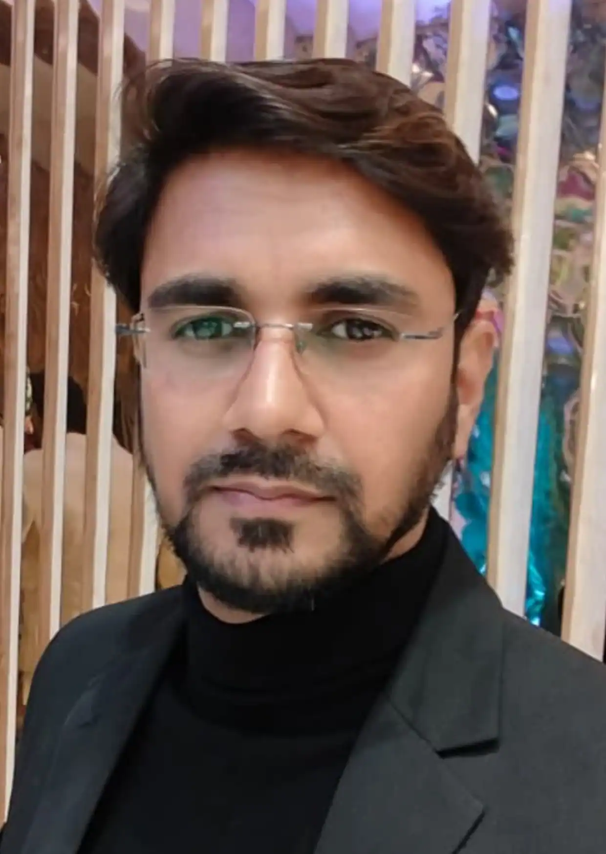 Professional portrait of Neal Bhai, a market analyst and financial expert, wearing a black suit and glasses, posing confidently indoors with a modern wooden panel background.